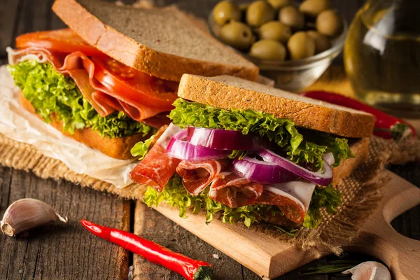 Close-up photo of a club sandwich. Sandwich with meet, prosciutto, salami, salad, vegetables, lettuce, tomato, onion and mustard on a fresh sliced rye bread on wooden background. Olives background.