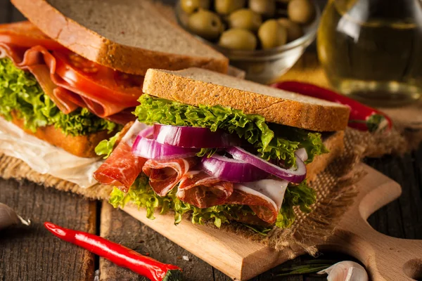 Close-up photo of a club sandwich. Sandwich with meet, prosciutto, salami, salad, vegetables, lettuce, tomato, onion and mustard on a fresh sliced rye bread on wooden background. Olives background.