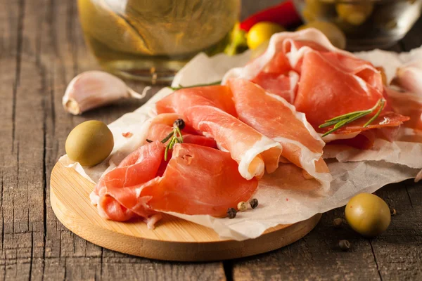 Närbild av tunna skivor prosciutto med blandade oliver och paprika på trä skärbräda — Stockfoto