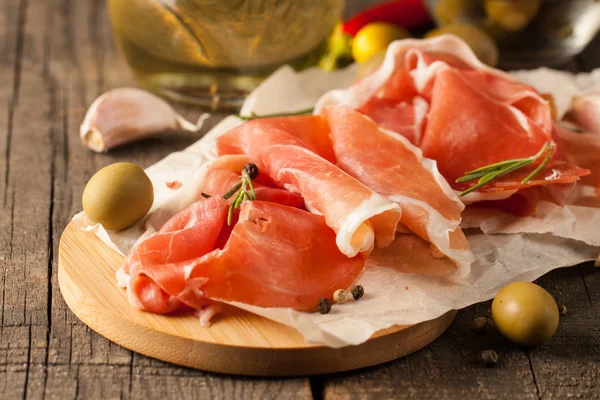 Närbild av tunna skivor prosciutto med blandade oliver och paprika på trä skärbräda — Stockfoto