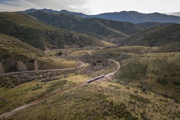 Aerial view of a train travelling along a track next to a road that winds its way along a valley in a natural park in the Balagne region of Corsica
