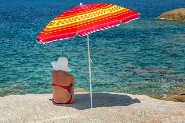 Woman in red bikini and white hat under parasol looking out to s