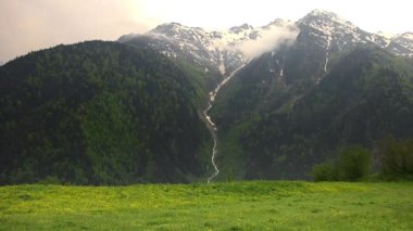 Sarı Çiçekli Görkemli Çayır 'daki Karlı Dağ Tepeleri. Sarı çiçekli çayırda bulutlu dağ iklimi. Dağlık alanlarda iklim değişkeni hareketleri. Dağlardaki derin vadiler. 4K