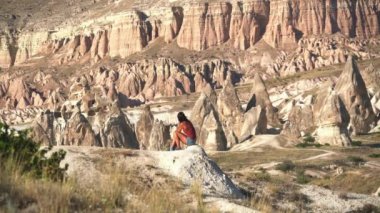 Göz kamaştırıcı kırmızı kumtaşı hoodoos manzarasında oturan bir kadın. Ulusal park büyücüsü genç kız çadır çadırı, kaya perisi baca piramit kanyon kapadokya doğa insanlar kurak drenaj havzası badland turistik tortu 4K