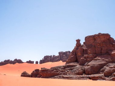 Sahra Çölü 'ndeki Kızıl Tadrart' ın kaya oluşumları ve altın tepecikleri. Berrak bir gökyüzünün altında çarpıcı bir doğal manzara. Sahra Çölü, Cezayir 'de gün doğumunda kum tepeleri ve kaya oluşumlarıyla Kızıl Tadrart' ın manzarası.