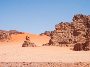 Sahra Çölü 'ndeki Kızıl Tadrart' ın kaya oluşumları ve altın tepecikleri. Berrak bir gökyüzünün altında çarpıcı bir doğal manzara. Sahra Çölü, Cezayir 'de gün doğumunda kum tepeleri ve kaya oluşumlarıyla Kızıl Tadrart' ın manzarası.