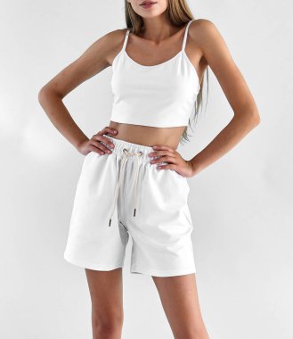 Genç kadın beyaz yaz kıyafetleri içinde duruyor. Beyaz kolsuz gömlek ve beyaz şort giyen bir kızın izole edilmiş portresi..