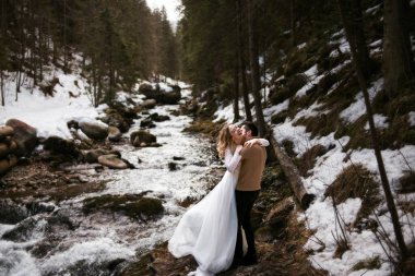 Damat ve gelin çifti, su deresi yakınlarındaki kış ormanında sarılıyorlar.