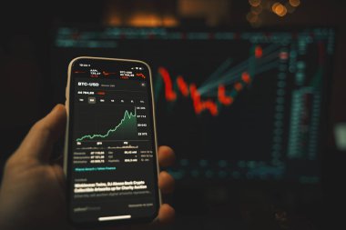 Elinde borsa uygulaması ve arka planda bulanık ekranı olan modern bir akıllı telefon var.