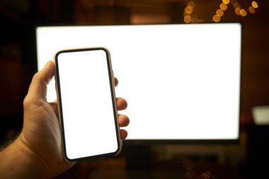 Arka planda bilgisayar monitörü olan modern akıllı telefonun el modeli. Boş ekran tasarım şablonu