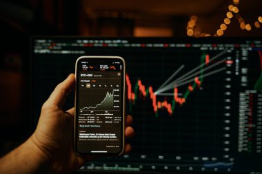 Elinde borsa uygulaması ve arka planda bulanık ekranı olan modern bir akıllı telefon var.