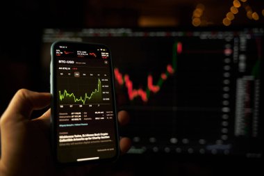 Elinde borsa uygulaması ve arka planda bulanık ekranı olan modern bir akıllı telefon var.