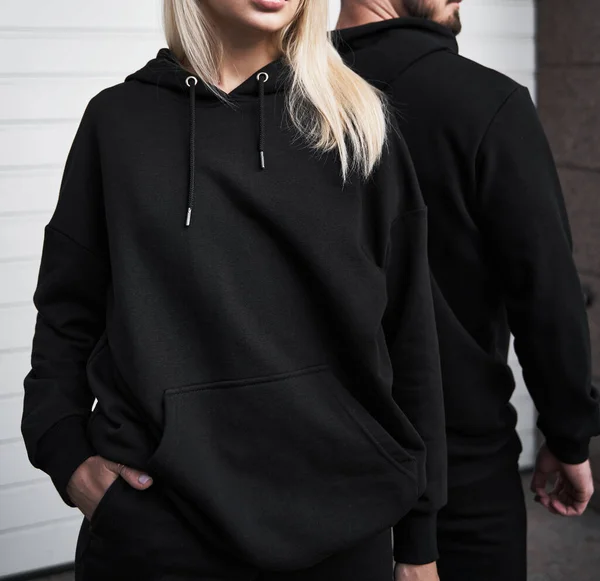 Woman stands in black no logo hoodie with bearded man behind her. Clothes branding mockup. Design template for casual sportswear
