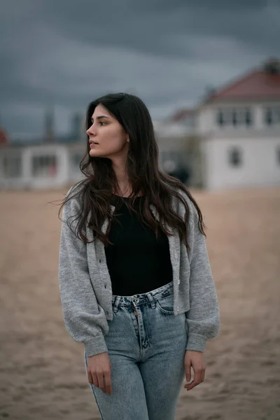 Teanager with long dark hair on the beach during cloudy evening. Brunette girl wears clothes near the sea during cold evening. Isolated portrait of lonely woman.