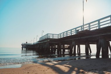 Deniz kıyısındaki güzel ahşap iskele. Yaz boyunca güçlü güneş ışığı. Ahşap zemini olan ve Baltık Denizi üzerinde beyaz tırabzanlara boyanmış gezinme yeri..