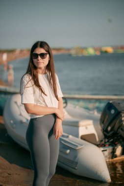 Kadın güneş gözlüğü takıyor ve sahilde tekneye karşı duruyor.