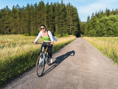 Çevresi yemyeşil olan kır yolunda bisiklet süren bir kadın.