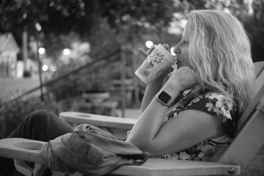 Tek kullanımlık bardaktan kahve içen bir kadın açık havada tek renkli bir ortamda oturuyor.