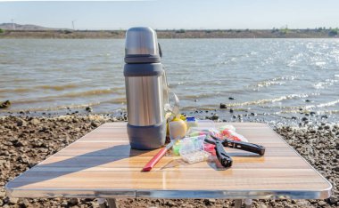 Tahta bir termosun üzerinde bir çoklu alet, bir bıçak, bir kutu olta takımı ve meyve var.