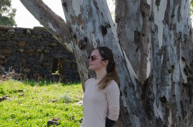 Güneş gözlüklü kız model, arkası büyük bir ağaca dönük duruyor.