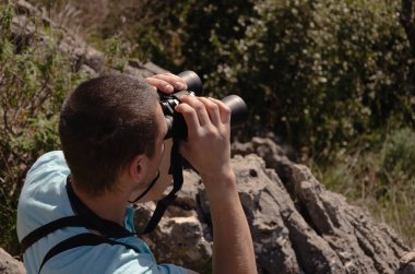 Genç bir adam dağlarda dürbünle manzaraya bakıyor.