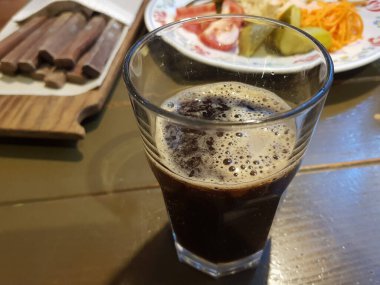 Öndeki masada üzerinde koyu kvass olan bir cam bardak.