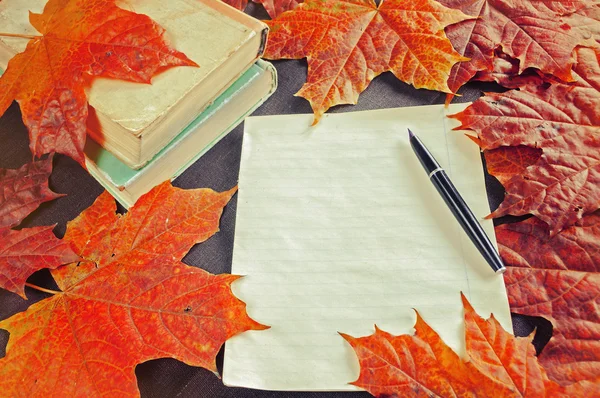 Autumn still life in vintage tones - old books near yellowed sheet of paper and old vintage ink pen on the table among yellow maple leaves.