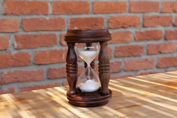 An hourglass made of brown wood. There is a brick wall in the background.