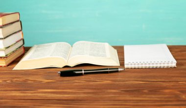 Masanın üzerinde açık bir kitap, defter, mavi arka planda bir kalem..