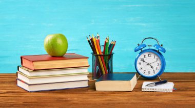 Bir yığın kitap, bir çalar saat, açık bir kitap, renkli kalemler, masada yeşil bir elma. Boşluğu kopyala.