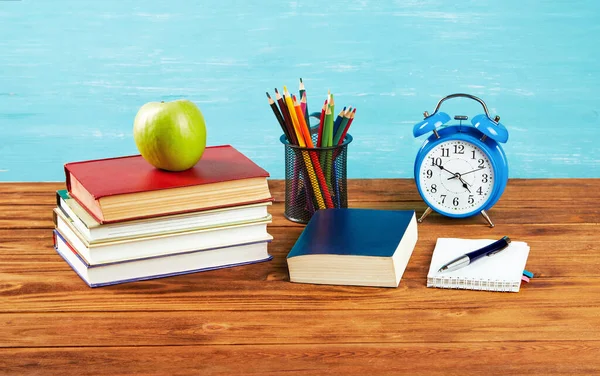  Bir yığın kitap, bir çalar saat, açık bir kitap, renkli kalemler, masada yeşil bir elma. Boşluğu kopyala.