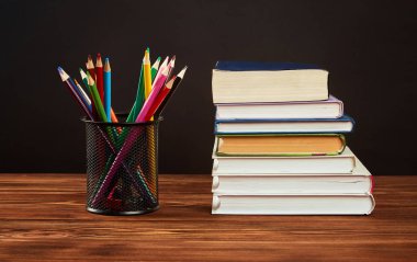 Renkli kalemler, siyah arka planda tahta bir masanın üzerinde bir yığın kitap..