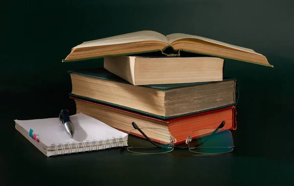 A stack of books, glasses, a notebook and a pen on a dark green background.