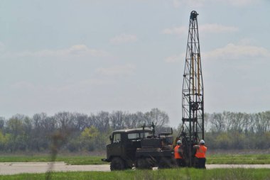 Jeodezi çalışmaları yerde yapılıyor, özel bir araç, arazi aracı ve işçiler görüyoruz.