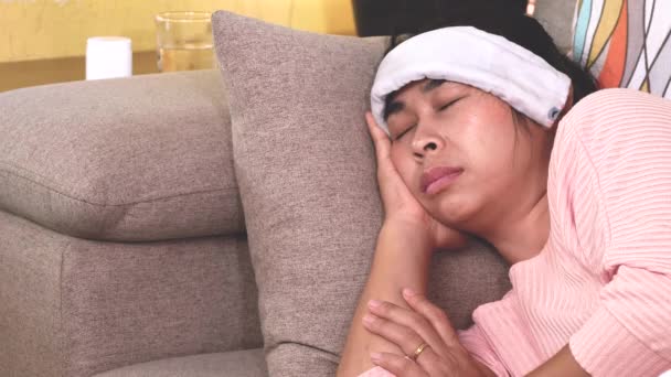 病気の女性は自宅のソファの上に横たわる喉の痛みと発熱頭痛を緩和するために彼女の額にぬれたタオルを置きます — ストック動画