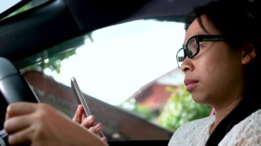 Genç bir kadın tek eliyle araba kullanırken yola bakıyor ve navigasyon kullanıyor ya da cep telefonlarından kitap okuyor..