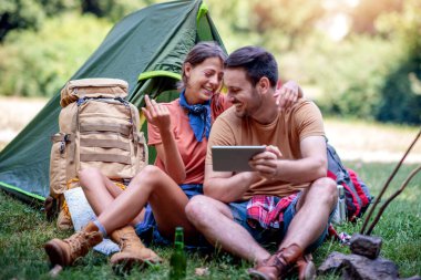 Çadırın önünde oturup tablet kullanan bir çift. Ormanda kamp yapmak..