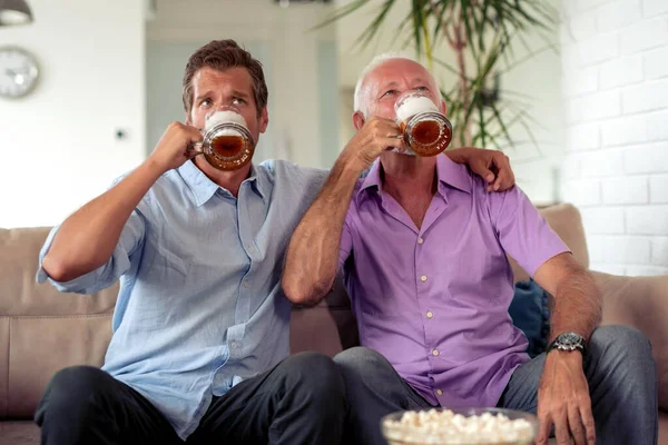 Father and son drink beers and watching football game at home.