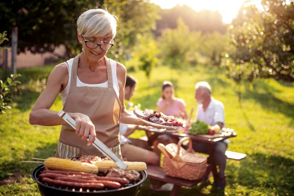 Food, people and family time concept.Senior woman cooking meat on barbecue grill at summer party.
