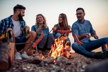 Bir grup arkadaş gün batımında ateşin etrafında oturmuş sosis pişiriyor ve kumsalda harika vakit geçiriyorlar.. 