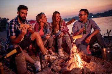 Bir grup arkadaş gün batımında ateşin etrafında oturmuş sosis pişiriyor ve kumsalda harika vakit geçiriyorlar.. 