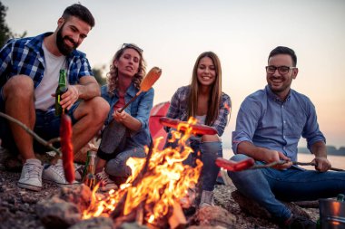 Bir grup arkadaş gün batımında ateşin etrafında oturmuş sosis pişiriyor ve kumsalda harika vakit geçiriyorlar.. 
