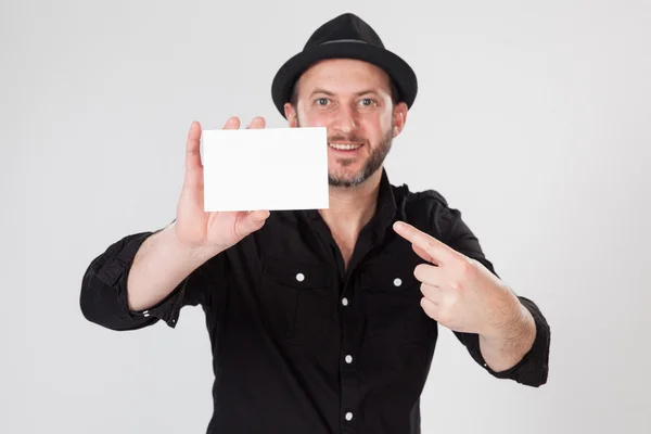 Χαμογελαστός άνθρωπος στη μαύρη μπλούζα και μαύρο καπέλο κρατώντας και δείχνοντας κενή κάρτα — Φωτογραφία Αρχείου