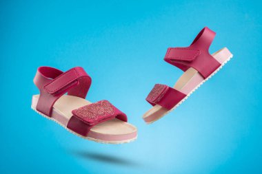Bir çift şık pembe çocuk sandaleti ve parlak mavi arka planda yüzen parlak detaylar. Son moda yaz ayakkabıları yaratıcı ve minimalist bir kompozisyonda çekildi..