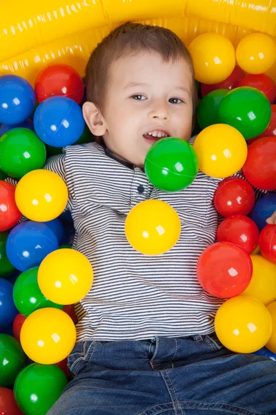 Маленький улыбающийся мальчик, играющий в разноцветные мячи на детской площадке — стоковое фото