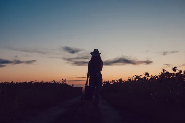 Bir kovboy şapkası ayakta ayçiçeği alan de yol bavulla kız. Yardım bekliyor. Günbatımı.