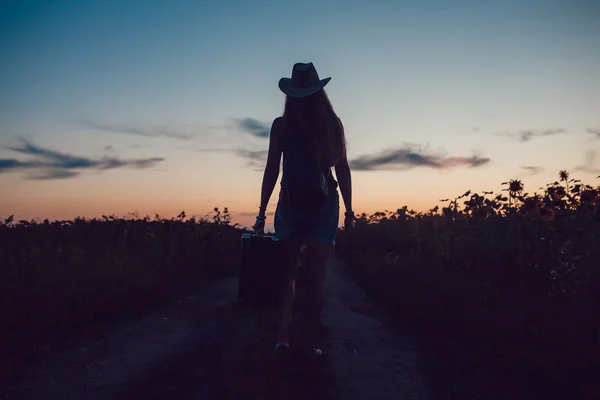 Κορίτσι σε μια μόνιμη καπέλο με μια βαλίτσα στο δρόμο στο πεδίο ηλιέλαιο. Αναμονή για βοήθεια. Ηλιοβασίλεμα. — Φωτογραφία Αρχείου