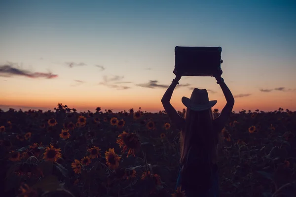 Menina com um chapéu de cowboy de pé com uma mala na estrada no campo de girassol. À espera de ajuda. Pôr do sol . — Fotografia de Stock