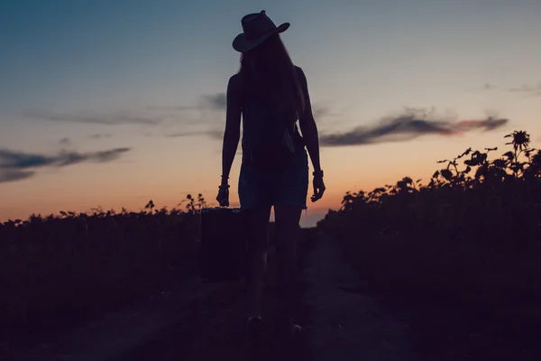 Mädchen mit Cowboyhut steht mit einem Koffer auf der Straße im Sonnenblumenfeld. Warten auf Hilfe. Sonnenuntergang. — Stockfoto