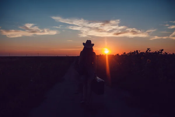 Girl in a cowboy hat standing with a suitcase on the road in the sunflower field. Waiting for help. Sunset. — Stock Photo, Image